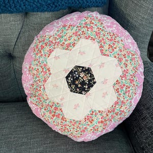 May include: A round pink and white floral patterned cushion with a quilted star design in the centre. The cushion is sitting on a grey sofa.