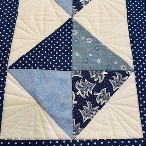 May include: A quilted table runner with a geometric design. The runner features white, light blue, and navy blue fabrics. The navy blue fabric has a white polka dot pattern. The white sections have a stitched fan design.