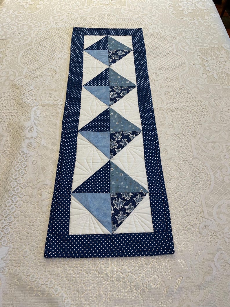 May include: A rectangular quilted table runner with a navy blue polka dot border. The centre features alternating white and blue diamond shapes, with various blue patterned fabrics. The runner is likely handmade and adds a decorative touch to a table.