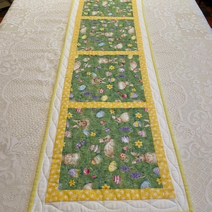 May include: A rectangular Easter-themed table runner. The center features a green fabric with bunnies, Easter eggs, and flowers. It is bordered by a yellow floral pattern and a white quilted edge. The runner is approximately 12 inches wide.