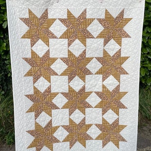 May include: A white quilt with a geometric pattern of brown stars. The quilt is hanging on a clothesline.