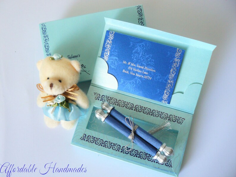 May include: A light blue invitation box with a white teddy bear wearing a blue dress and a blue and silver scroll inside. The box has a blue and silver design with the text "Fuliana's" on the front.