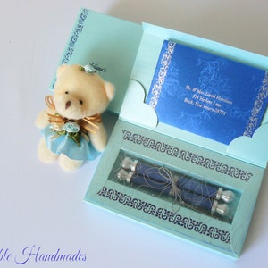May include: A light blue gift box with a teddy bear on top. The box is decorated with silver scrollwork and contains two rolled up scrolls with silver embellishments. The box is open and the scrolls are visible. The box is addressed to Mr. & Mrs. David Hamilton, 574 Herlies Lane, Brick, New Jersey 08724.