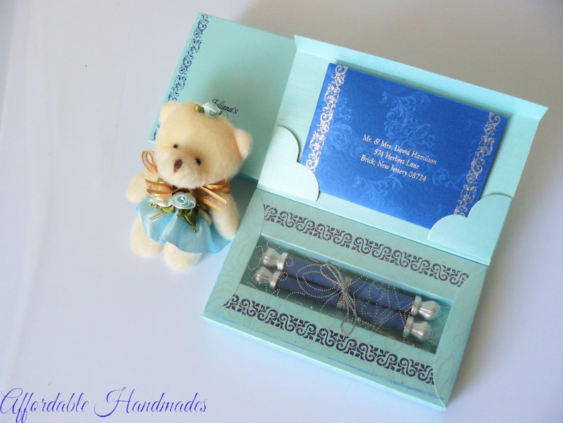 May include: A light blue gift box with a teddy bear on top. The box is decorated with silver scrollwork and contains two rolled up scrolls with silver embellishments. The box is open and the scrolls are visible. The box is addressed to Mr. & Mrs. David Hamilton, 574 Herlies Lane, Brick, New Jersey 08724.