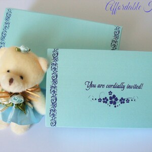 May include: A light blue invitation card with a white teddy bear wearing a blue dress and a gold bow. The card has a blue border with a floral design and the text "You are cordially invited!"