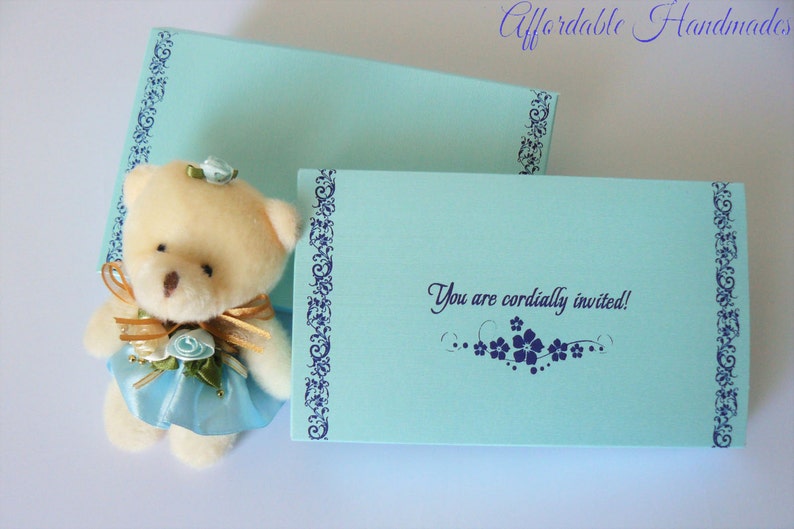 May include: A light blue invitation card with a white teddy bear wearing a blue dress and a gold bow. The card has a blue border with a floral design and the text "You are cordially invited!"