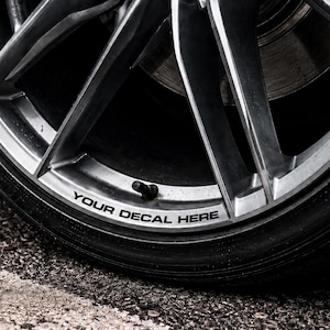 Könnte beinhalten: Nahaufnahme eines silbernen Leichtmetall-Autorads mit einem schwarzen Reifen. Die Felge hat ein Mehrspeichen-Design. Der Text "YOUR DECAL HERE" ist in Schwarz auf die Felge gedruckt. Das Rad befindet sich auf einer dunklen, strukturierten Oberfläche.