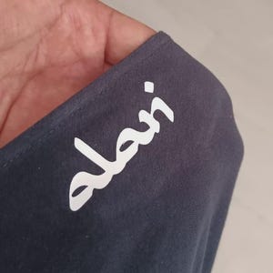 May include: A close-up of a dark gray fabric with the white word "alan" printed on it. The fabric is held up, showing its texture and the crisp white lettering. The background is a light gray surface.