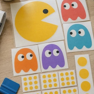 Op de afbeelding: Een verzameling vinylstickers met een grote gele Pac-Man-figuur, vier geesten in rood, blauw, oranje en paars, en meerdere kleine gele stippen. Een blauwe plastic rakel is inbegrepen voor het aanbrengen van de zelfklevende stickers.