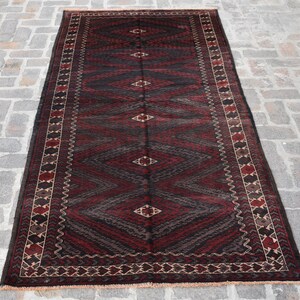 Può includere: Un tappeto persiano tradizionale con uno schema di colori rosso scuro e nero. Il tappeto presenta un motivo geometrico con forme a diamante e dettagli intricati sul bordo.