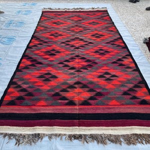 May include: A rectangular rug with a geometric diamond pattern in shades of red, black, and gray. The rug has a black border and fringed edges. The rug is displayed outdoors on a light-colored surface.