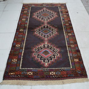 Pode incluir: Um tapete castanho escuro com um padrão geométrico em tons de vermelho, azul e branco. O tapete tem uma borda de franjas.