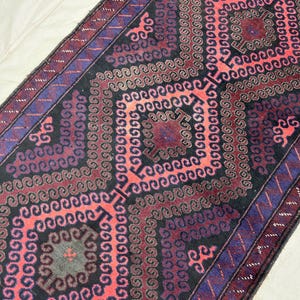 Pode incluir: Um tapete retangular com um padrão geométrico central em tons de vermelho, preto e roxo. O design apresenta formas de diamante repetidas e motivos em espiral. A borda é roxa escura com um detalhe azul.