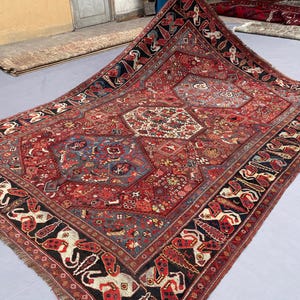 Op de afbeelding: Een groot rechthoekig vloerkleed met een dieprood veld en ingewikkelde geometrische en florale patronen in blauw, crème en zwart. De rand toont een herhalend motief van gestileerde dierenfiguren tegen een donkere achtergrond.