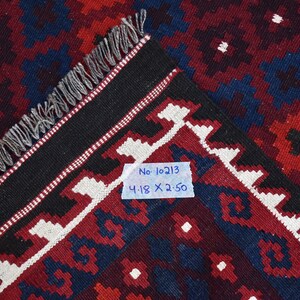 May include: Close-up of a red, blue, and white patterned rug with black fringe. A white tag with the text "No. 10213 4.18 X 2.50" is attached to the rug.