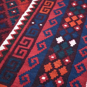 May include: A close-up of a red, blue, and white patterned rug with intricate geometric designs. The rug has a traditional, hand-woven look.