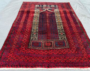 May include: A rectangular prayer rug featuring a deep red and dark blue color palette. The central field displays a mihrab arch design with intricate geometric patterns and borders. The rug is finished with white fringe along the short edges.