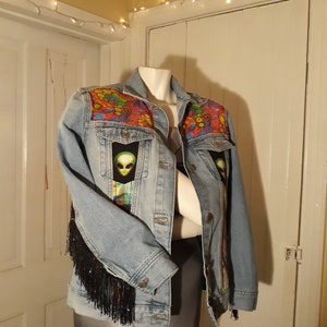 May include: A light blue denim jacket with black fringe on the sleeves. The jacket has colorful patches with alien faces on the front.
