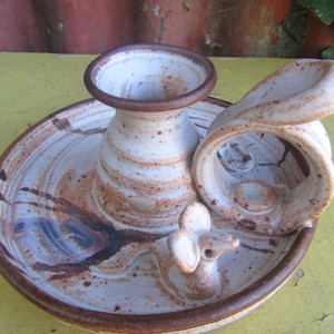 May include: A ceramic candle holder featuring a circular base with a central cup and a curved handle. The piece is finished in a speckled off-white glaze with brown accents and includes a small sculpted mouse figure attached to the base.