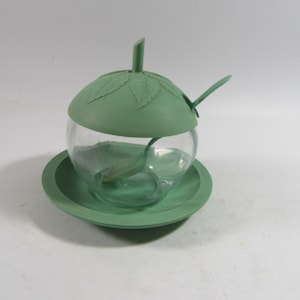 May include: A green, apple-shaped sugar bowl with a clear glass body and a matching lid and spoon. The lid is decorated with leaf details. The bowl sits on a round, green saucer. The spoon is inside the bowl.