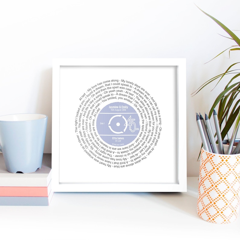 May include: A white framed print of a light blue vinyl record with the song "At Last" by Etta James. The lyrics are printed in a circle around the record. The record is labeled with "Side 1" and "45" in the center. The date "23 August 2018" and the names "Jasmine & Emily" are printed above the record.