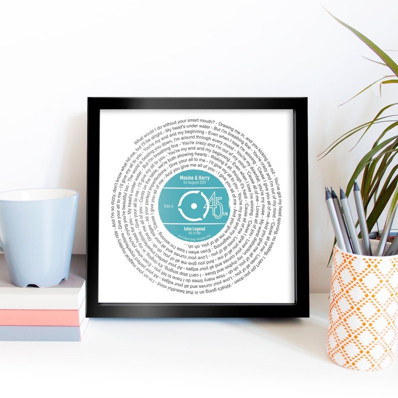 May include: A framed print of a vinyl record with the lyrics to the song "All of Me" by John Legend. The record is blue and white and has the names Maxine & Harry and the date 1st August 2019 printed on it.