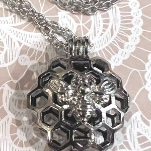 May include: A silver-toned pendant necklace featuring a circular honeycomb-patterned cage design. A detailed bee figure is centered on the front of the honeycomb structure. The pendant is attached to a matching silver-toned link chain necklace.