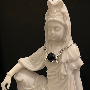 May include: A white statue of a seated figure wearing a silver necklace with a black stone pendant. The necklace features crescent moon accents on either side of the central stone. The statue is set against a dark background.