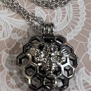 May include: Silver-toned necklace featuring a honeycomb pendant with a bee design. The pendant is detailed with hexagonal cells and a central bee figure. The chain is a delicate, linked style, suitable for everyday wear.