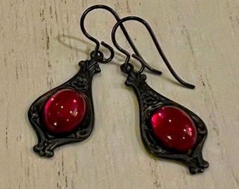 May include: A pair of dark metal earrings with a teardrop shape. Each earring features a vibrant red oval-shaped stone set within an ornate frame. The earrings have a hook closure.