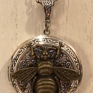 May include: Silver-tone locket necklace with a detailed bronze-coloured bee pendant. The round locket features an ornate floral design around the bee. The necklace has a chain with a decorative bail.