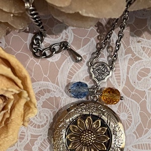 May include: Silver-tone sunflower locket necklace with a gold-tone sunflower design. The pendant has a decorative border and is suspended from a chain with blue and yellow bead accents. The necklace is displayed on a pink lace background.