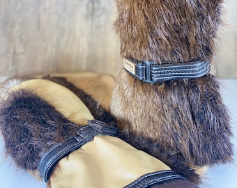 May include: A pair of brown fur mittens with leather straps and brown trim. The mittens have a tan leather palm and a brown fur cuff.
