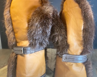 May include: A pair of brown leather mittens with fur trim. The mittens have a brown leather strap with a white stitch detail.