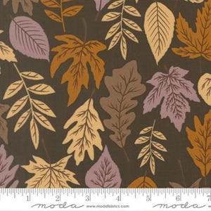 May include: A dark brown fabric featuring a repeating pattern of autumn leaves in shades of tan, orange, and muted purple. A white ruler is visible at the bottom edge, showing measurements in inches, with the text moda and modafabrics.com printed.
