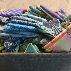 May include: A gray fabric storage basket filled with a variety of colorful fabric scraps. The scraps are folded and stacked, and they include patterns of paisley, floral, and geometric designs. The colors include blue, green, pink, and white.