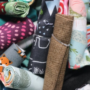 May include: A close-up of a pile of fabric scraps in various colors and patterns, including a pink and white polka dot fabric, a blue and white floral fabric, and a brown fabric with a textured surface.