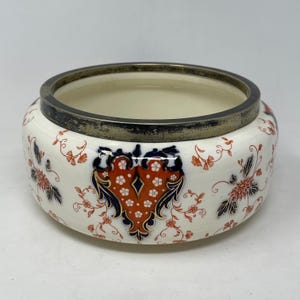 May include: A white ceramic bowl with a silver rim and a red, blue, and gold floral design. The bowl is decorated with a central floral motif and smaller floral patterns around the rim.