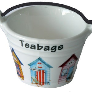 May include: White ceramic tea bag holder shaped like a bucket with a black handle. The bucket has a colourful beach hut design and the word "Teabags" printed on the side.