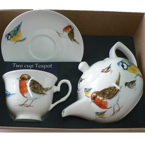 May include: A two-cup teapot and teacup set with a saucer. The set features a white background with a colourful bird design. The teapot and teacup have a robin and other birds on them. The saucer has a bluebird on it. The set is packaged in a brown box.
