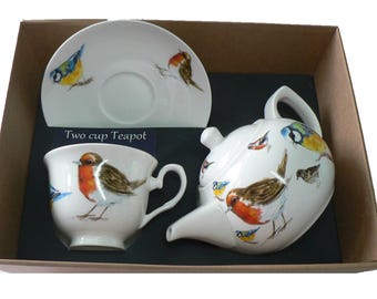 May include: A two-cup teapot and teacup set with a saucer. The set features a white background with a colorful bird design. The teapot and teacup have a robin and other birds on them. The saucer has a bluebird on it. The set is packaged in a brown box.