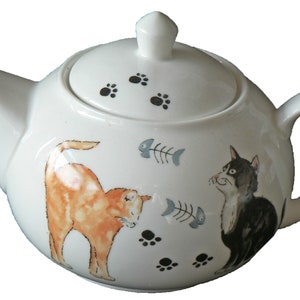 May include: White ceramic teapot with a lid, spout, and handle. The teapot is decorated with illustrations of an orange cat, a black and white cat, fish bones, and paw prints.