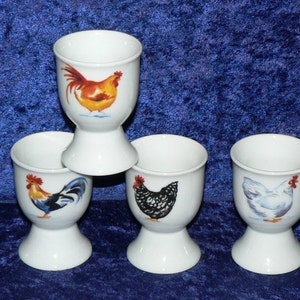 May include: Four white ceramic egg cups with different chicken designs. The egg cups have a white base and a white rim. The designs are a red rooster, a black and white hen, a white hen, and a yellow rooster.