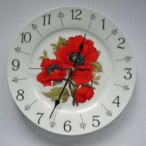 May include: A white porcelain clock with black Roman numeral hour markers and black hands. The clock face features a floral design with red poppies and green leaves.