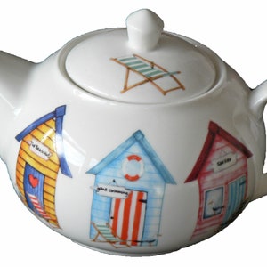May include: White ceramic teapot with a lid, decorated with colourful beach hut illustrations. The lid features a beach chair design. The teapot has a curved handle and spout, perfect for serving tea.