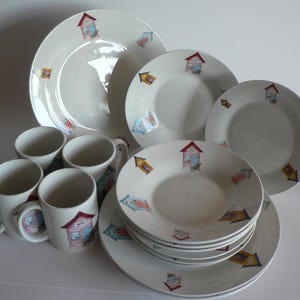 May include: A set of white ceramic mugs and plates featuring a beach house design. The mugs and plates have a colourful illustration of beach houses with red, blue and yellow accents.