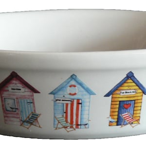 May include: White ceramic oval bowl with a beach hut design. The bowl features three beach huts with different colours and designs, including a red and blue hut, a blue and white striped hut, and a yellow hut with a red heart.