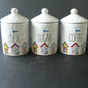 May include: Three white ceramic canisters with lids, each featuring a textured surface, a blue seagull illustration, and a row of colourful beach huts at the base. The canisters are labelled with black text reading TEA, SUGAR, and COFFEE respectively.