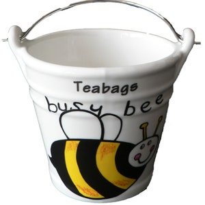 May include: White ceramic tea bag holder shaped like a bucket with a silver handle. The bucket has a black and yellow bee illustration with the text "Teabags busy bee".