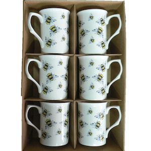 May include: Set of six white ceramic mugs with a bee pattern. The mugs have black handles and are arranged in a brown box.